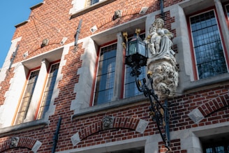 A historic building facade with red brick walls and stone accents. The windows are tall and framed with white stone. A detailed stone sculpture featuring a person and ornate carvings prominently decorates the building, positioned near a black wrought-iron lantern.