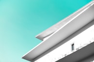A minimalist architectural design featuring clean lines and geometric shapes, with a bright turquoise sky in the background. The building facade is white with sharp angles and one visible window.
