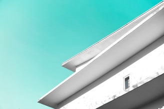 A minimalist architectural design featuring clean lines and geometric shapes, with a bright turquoise sky in the background. The building facade is white with sharp angles and one visible window.