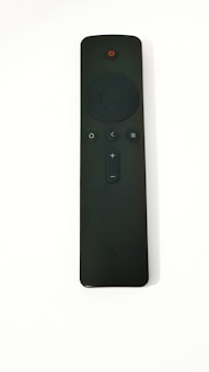 A black remote control with several buttons, including a circular directional pad, power button at the top, and volume control. The surface appears matte with a minimalist design.