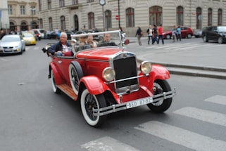 A vintage red convertible car with large chrome headlights and white-wall tires drives down a city street. The car carries several people who appear to be enjoying a leisurely ride. The background shows a historic urban setting with old-style buildings and other cars. Pedestrians can be seen walking on the sidewalk, adding to the lively city atmosphere.