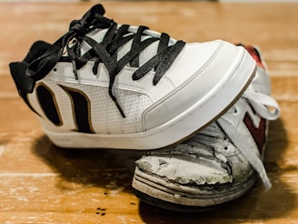 A new, clean sneaker with black and white colors is placed on top of a very worn and damaged sneaker. The worn sneaker shows significant signs of wear with visible scuffs, dirt, and peeling material. Both shoes are positioned on a wooden surface with visible wear and grain.