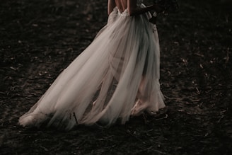 A bride in an elegant, flowing white gown is walking on a dark, earthy ground. The dress features delicate tulle fabric that drapes gracefully to the floor, creating a sense of movement and grace.