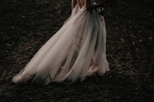 A bride in an elegant, flowing white gown is walking on a dark, earthy ground. The dress features delicate tulle fabric that drapes gracefully to the floor, creating a sense of movement and grace.