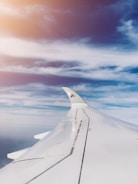 A view from an airplane window showing the aircraft's wing and a partially visible vertical stabilizer with a distinctive logo. The sky is filled with fluffy, wispy clouds beneath a bright blue sky with a soft glowing light from the sun at the top left corner.