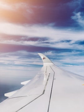 A view from an airplane window showing the aircraft's wing and a partially visible vertical stabilizer with a distinctive logo. The sky is filled with fluffy, wispy clouds beneath a bright blue sky with a soft glowing light from the sun at the top left corner.