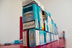 A stack of various cigarette packs, prominently featuring the Camel brand, arranged on a red surface. The packs display warning labels in different languages. The image has a shallow depth of field, with the focus on the nearer packs and a blurred background.