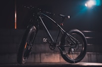 A mountain bike with thick tires is positioned against a dark, moody background with a spotlight illuminating it from the side, creating dramatic shadows. The bike's details, including the handlebars and pedal, are partially visible in the subdued lighting.