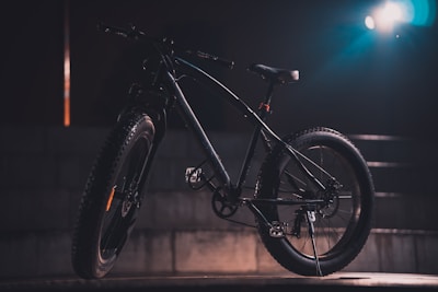 A mountain bike with thick tires is positioned against a dark, moody background with a spotlight illuminating it from the side, creating dramatic shadows. The bike's details, including the handlebars and pedal, are partially visible in the subdued lighting.