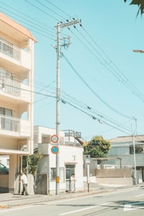 A quiet urban street with a light-colored building on the left and a residential area in the background. Power lines stretch across the clear blue sky, intersecting with a tall utility pole. There are signs along the street, including a round speed limit sign and a no parking sign. Two individuals are visible, with one standing near the building and the other walking.
