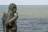 A bronze statue of a bearded man in historical attire holding a staff, with the vast ocean in the background. The statue is positioned sideways, looking towards the sea.