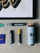 A collection of art supplies is displayed on a light gray surface. The items include a framed artwork featuring stylized text in black and gold, several markers, a spray paint can, and a small purple booklet. The markers are of different sizes, with one having a yellow cap. A small sticker with colorful text is also visible.