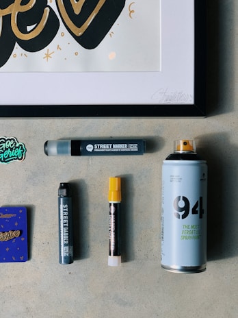A collection of art supplies is displayed on a light gray surface. The items include a framed artwork featuring stylized text in black and gold, several markers, a spray paint can, and a small purple booklet. The markers are of different sizes, with one having a yellow cap. A small sticker with colorful text is also visible.