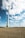 Tall wind turbines rise above an open field, under a sky filled with scattered clouds. The foreground shows a textured earth surface, likely a field or farmland. Several turbines are visible, with the closest one towering over the landscape, suggesting a focus on renewable energy.