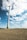 Tall wind turbines rise above an open field, under a sky filled with scattered clouds. The foreground shows a textured earth surface, likely a field or farmland. Several turbines are visible, with the closest one towering over the landscape, suggesting a focus on renewable energy.