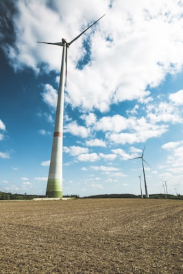Tall wind turbines rise above an open field, under a sky filled with scattered clouds. The foreground shows a textured earth surface, likely a field or farmland. Several turbines are visible, with the closest one towering over the landscape, suggesting a focus on renewable energy.