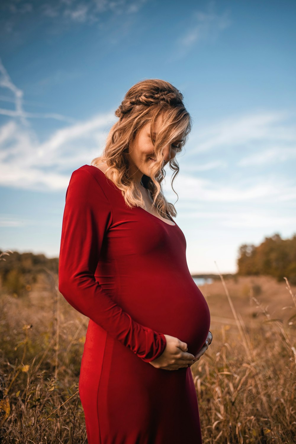 smiling pregnant woman wearing red scoop-neck long-sleeved dress touching her tummy while standing on brown field under white and blue sky during daytime