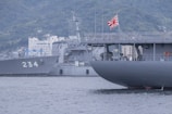 Two military ships are docked in a harbor with a mountainous background. One ship has a visible number 234 on its side, while another displays a prominent flag with a red and white rising sun design. Buildings and green hills are visible in the distance.