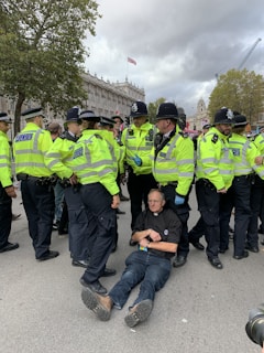 A group of police officers in neon green jackets surrounds a man sitting on the ground. The man appears calm and is wearing a clerical shirt. In the background, there is a building with ornate architecture and a partially cloudy sky.
