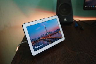 A digital smart display sits on a wooden desk, showing a vibrant cityscape with a tall tower and skyscrapers at dusk. The screen displays the time as 1:32 with a temperature of 29°C. In the background, a speaker and another screen are slightly visible.