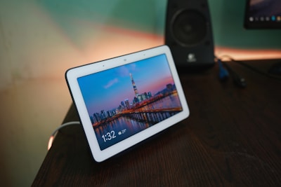 A digital smart display sits on a wooden desk, showing a vibrant cityscape with a tall tower and skyscrapers at dusk. The screen displays the time as 1:32 with a temperature of 29°C. In the background, a speaker and another screen are slightly visible.