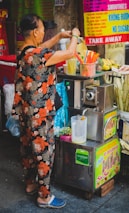 A person in floral attire operates a juice stand, with various colorful signs in the background advertising smoothies and sugarcane juice. On the stand, there are vibrant straws in a container, and a pitcher with a citrus juicer attached.