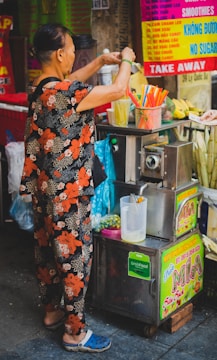 A person in floral attire operates a juice stand, with various colorful signs in the background advertising smoothies and sugarcane juice. On the stand, there are vibrant straws in a container, and a pitcher with a citrus juicer attached.