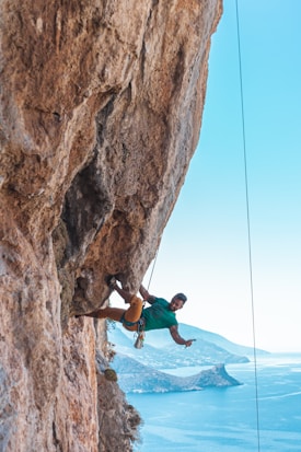 A rock climber wearing a green shirt and tan shorts is scaling a steep cliff face, with a picturesque ocean and mountainous landscape in the background. The climber is securely attached to a rope and appears to be in mid-movement, gripping the rock with one hand and stretching out his other arm for balance.