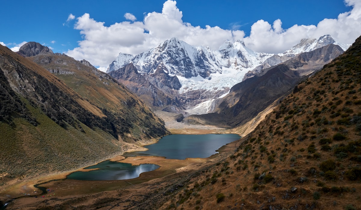 Turquoise glacial lake in a steep brown valley beneath snow-covered Andean peaks under a bright blue sky
