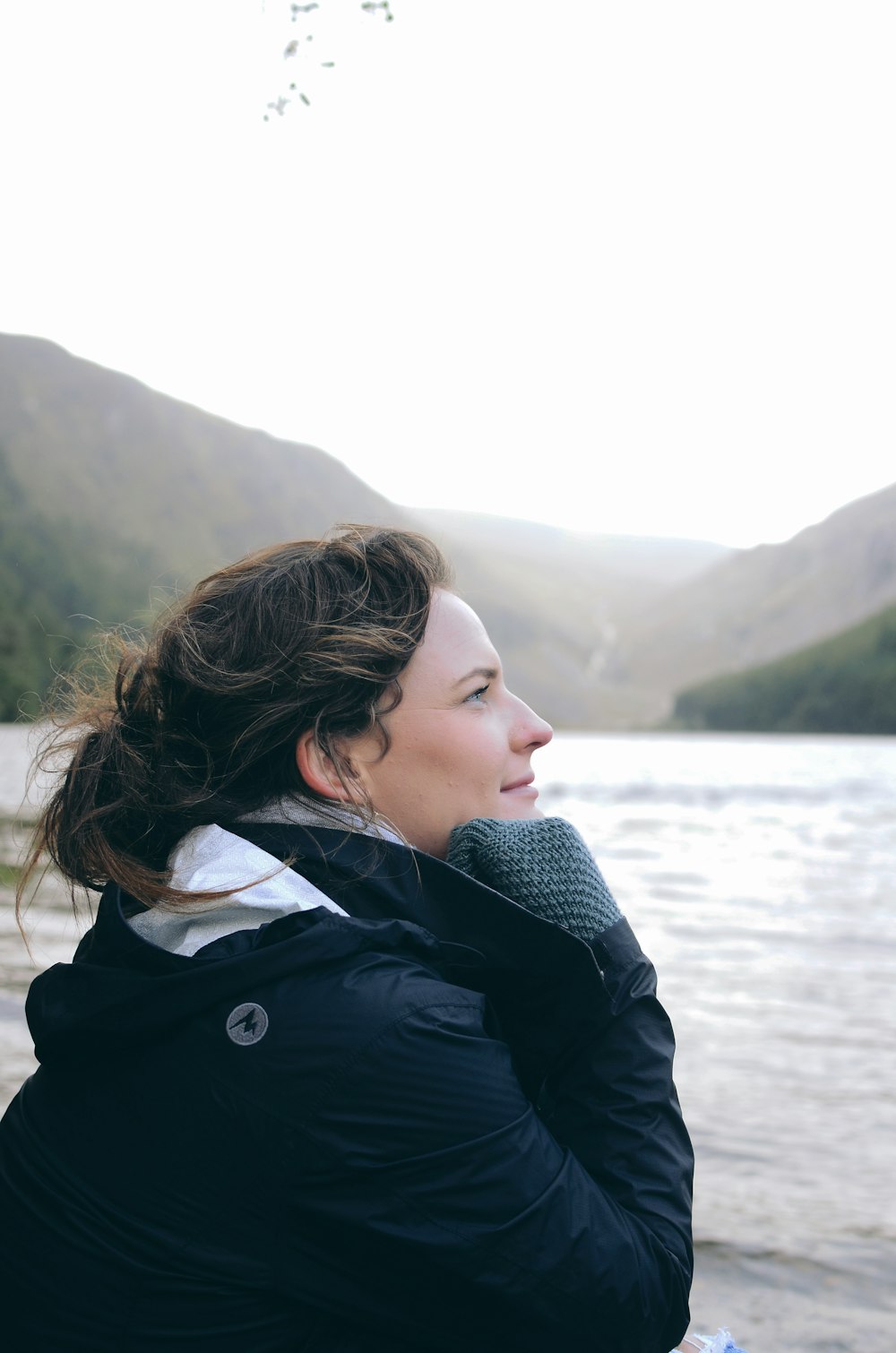 Mujer con chaqueta blanca y negra mirando hacia su lado izquierdo cerca del cuerpo de agua que ve la montaña durante el día