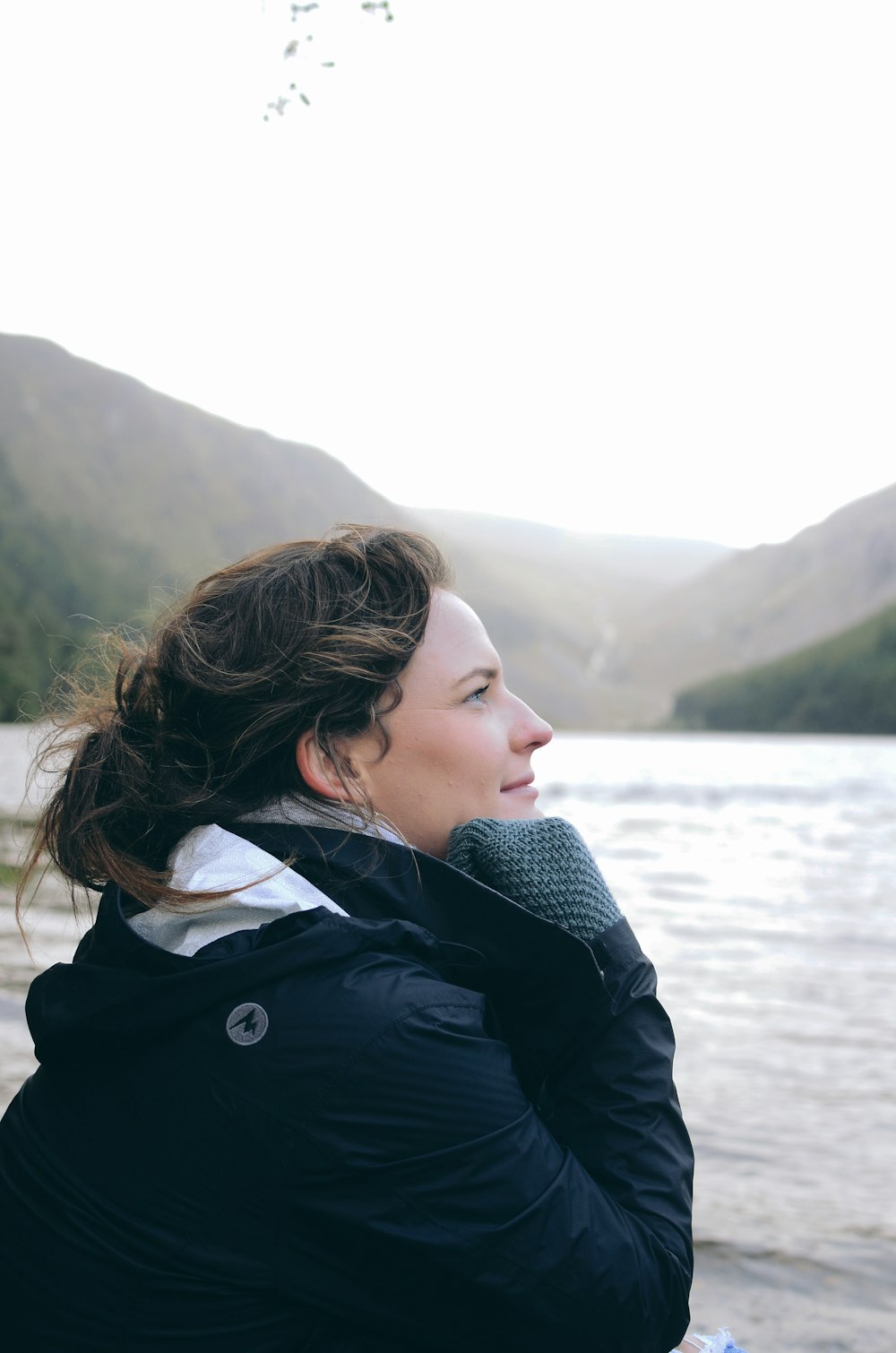 woman wearing black and white jacket facing her left side near body of water viewing mountain during daytime