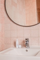 A bathroom sink with a chrome faucet is positioned in front of a pink tiled wall. On the sink counter, there is a white bottle labeled 'Curology' with an open cap placed beside it. A round mirror with a thin metal frame hangs above the sink.