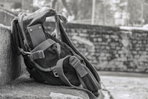 A rugged backpack rests against a stone wall, showcasing a sturdy design with reinforced straps and multiple compartments. The background features a blurred stone wall and foliage, suggesting an outdoor setting.