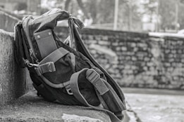 A rugged backpack rests against a stone wall, showcasing a sturdy design with reinforced straps and multiple compartments. The background features a blurred stone wall and foliage, suggesting an outdoor setting.
