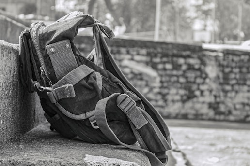 A rugged backpack rests against a stone wall, showcasing a sturdy design with reinforced straps and multiple compartments. The background features a blurred stone wall and foliage, suggesting an outdoor setting.