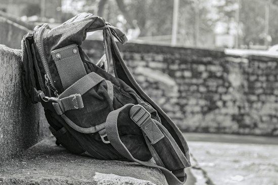 A rugged backpack rests against a stone wall, showcasing a sturdy design with reinforced straps and multiple compartments. The background features a blurred stone wall and foliage, suggesting an outdoor setting.
