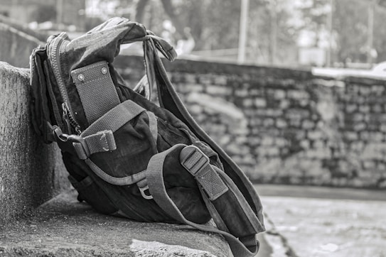 A rugged backpack rests against a stone wall, showcasing a sturdy design with reinforced straps and multiple compartments. The background features a blurred stone wall and foliage, suggesting an outdoor setting.