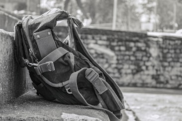 A rugged backpack rests against a stone wall, showcasing a sturdy design with reinforced straps and multiple compartments. The background features a blurred stone wall and foliage, suggesting an outdoor setting.
