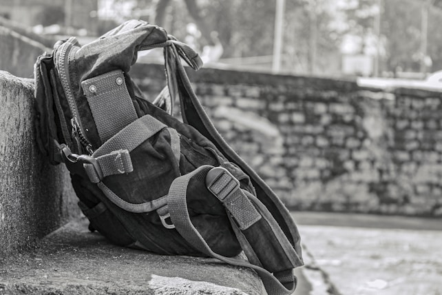 A rugged backpack rests against a stone wall, showcasing a sturdy design with reinforced straps and multiple compartments. The background features a blurred stone wall and foliage, suggesting an outdoor setting.