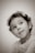 A sepia-toned portrait of a young child with curly hair looking upwards with a playful expression. The child is wearing a floral-patterned top and small earrings, set against a plain backdrop.