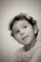 A sepia-toned portrait of a young child with curly hair looking upwards with a playful expression. The child is wearing a floral-patterned top and small earrings, set against a plain backdrop.