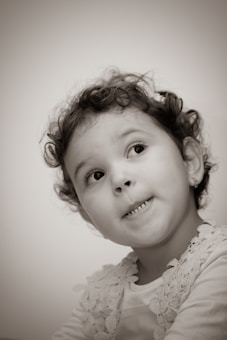 A sepia-toned portrait of a young child with curly hair looking upwards with a playful expression. The child is wearing a floral-patterned top and small earrings, set against a plain backdrop.