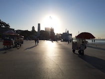 A coastal promenade during sunset featuring street vendors with colorful umbrellas. There is a large ship docked nearby, and people are walking and relaxing in the open space. The scene is urban with tall buildings and palm trees visible in the distance.