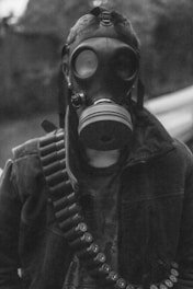 A person is wearing a gas mask and a jacket with a bandolier of shotgun shells across the chest. The mask covers the entire face, emphasizing a sense of anonymity or protection. The background is blurred, adding focus to the figure.