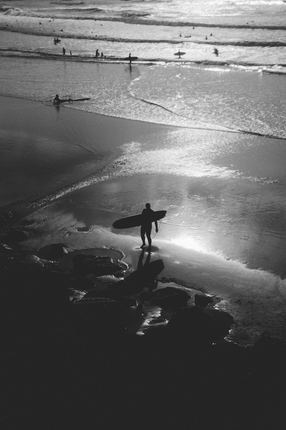 Graustufenfotografie einer Person, die ein Surfbrett am Meer trägt