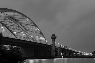 An arch bridge spans across a body of water with lights illuminating its structure. A control tower stands prominently by the bridge. The image is in black and white, enhancing the architectural details and reflections on the water.