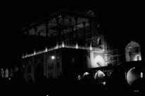 A large, intricately designed structure with arches and columns is illuminated at night. The architecture is highlighted by sparse lighting, creating dramatic contrasts and shadows. Part of the building appears to be under construction, with scaffolding visible.
