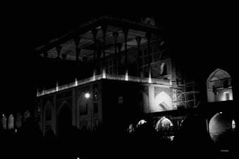 A large, intricately designed structure with arches and columns is illuminated at night. The architecture is highlighted by sparse lighting, creating dramatic contrasts and shadows. Part of the building appears to be under construction, with scaffolding visible.