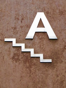 A metallic letter 'A' followed by a zigzag line resembling stairs, mounted on a textured brown wall.