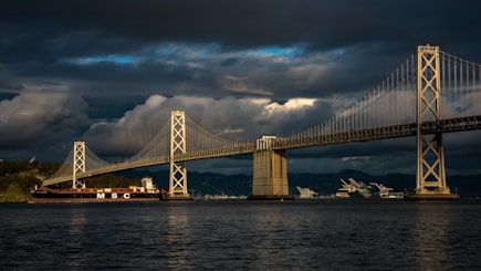 A large cargo ship with 'MSC' written on its side sails beneath a steel suspension bridge. Dark, dramatic clouds fill the sky, casting shadows on the water below, while sunlight breaks through, highlighting parts of the landscape and infrastructure. In the background, cranes and industrial structures are visible along the shoreline.